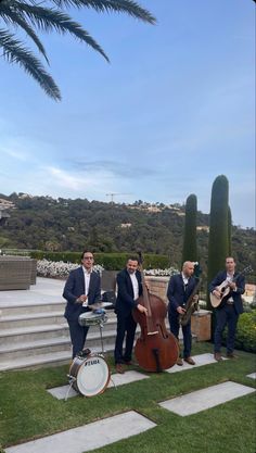 three men in suits are playing instruments on the steps near some trees and bushes, while another man is standing behind them