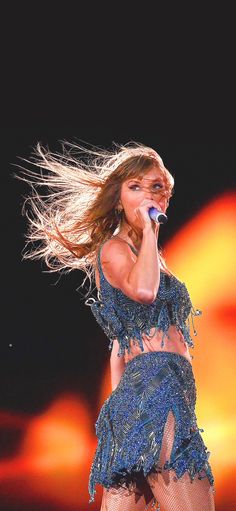 a woman in a blue dress on stage with her hair blowing in the wind and holding a microphone to her ear