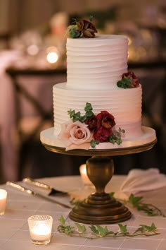 a white wedding cake with red flowers and greenery sits on a gold pedestal next to candles