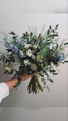 a person holding a bouquet of blue and white flowers with greenery in their hands