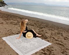 a woman sitting on top of a beach next to the ocean with a hat in her hand