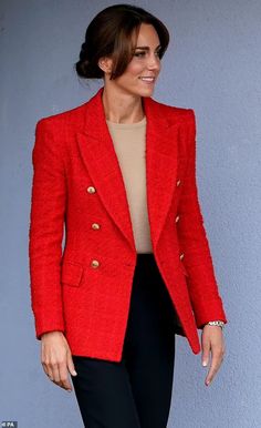 a woman in a red jacket and black pants smiles as she stands against a wall