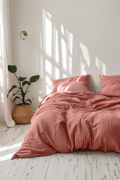 an unmade bed with pink sheets and pillows in a white room next to a potted plant