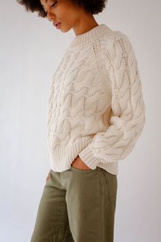 a woman wearing green pants and a white cable knit sweater, looking down at her stomach