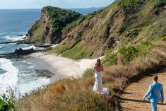 a bride and groom walking down a path to the beach with cliffs in the background