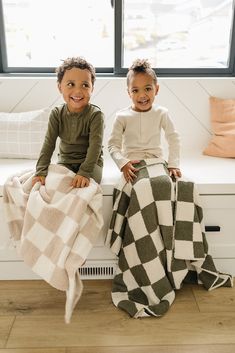 two children sitting on a window sill under a checkered blanket and smiling at the camera