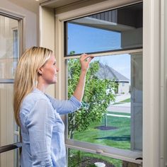 a woman standing in front of a window looking out at the grass and trees outside