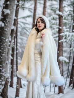 a woman in a white dress and fur coat standing in the snow with trees behind her