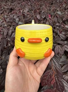 a hand holding a yellow ceramic cup with black eyes and an orange nose on it