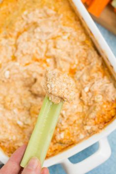 a hand holding a celery stick over a casserole dish with carrots in the background