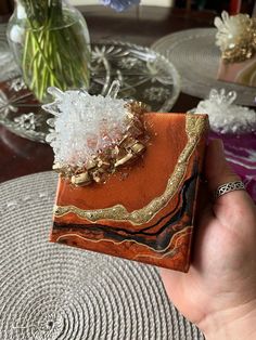 a hand holding an orange and gold box with some flowers in the vase behind it