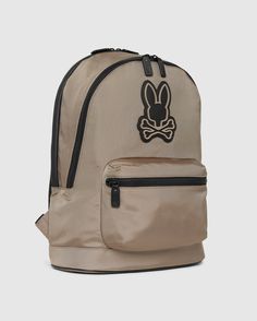 A tan MENS DOME BACKPACK - B6A628Z1BP by Psycho Bunny features a black bunny logo with crossbones on the front. It boasts a large main compartment, a padded laptop compartment, and a smaller front zippered pocket, all accented with black zippers and straps. The background is plain white. Daily Use Backpack With Logo Patch, Casual Everyday Backpack With Logo Patch, Streetwear Backpack With Functional Pockets, Nylon Backpack With Logo Patch, Streetwear Backpack With Zipper Closure, Modern Backpack With Logo Patch, Functional Backpack With Logo Patch, Streetwear Backpack With Pockets, Functional Everyday Backpack With Logo Patch