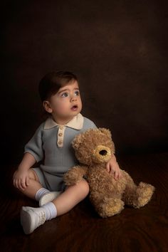 a baby sitting next to a teddy bear on a wooden floor in front of a dark background