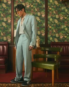 a man in a blue suit and sunglasses standing next to a green chair with a floral wallpaper behind him