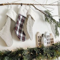 christmas stockings hung on a mantel decorated with greenery and pine cones, along with miniature houses