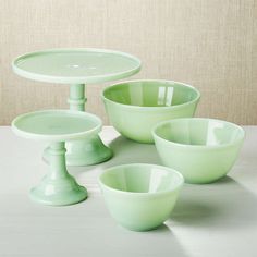 three green bowls and two cake plates on a white tableclothed surface with a beige wall in the background