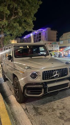 a silver mercedes g class is parked on the side of the road at night time