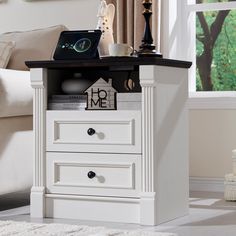 a white nightstand with two drawers and a clock on it's side table next to a couch