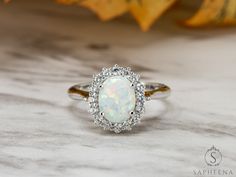 an opal and diamond ring sits on a marble surface with leaves in the background