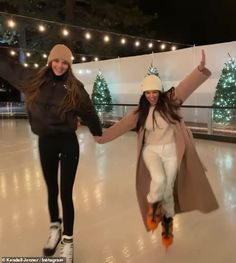 two women skating on an ice rink at night with christmas lights in the back ground