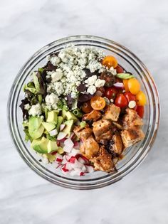 a glass bowl filled with different types of vegetables and meats on top of a marble table