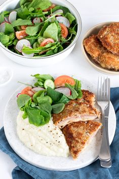 two plates with meat and vegetables on them next to a bowl of salad, fork and knife