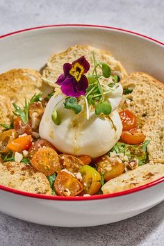 a bowl filled with bread and vegetables on top of a table next to a purple flower