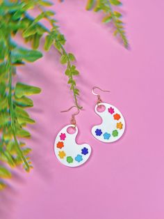 two pairs of white earrings with colorful flowers on them next to some green plant leaves