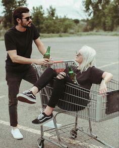 a woman sitting in a shopping cart with a man standing next to her holding a beer