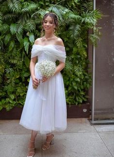 a woman standing in front of a green wall wearing a white dress and holding a bouquet