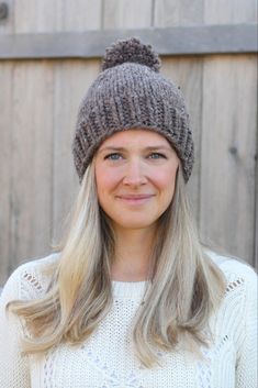 a woman with blonde hair wearing a knitted hat and smiling at the camera while standing in front of a wooden fence