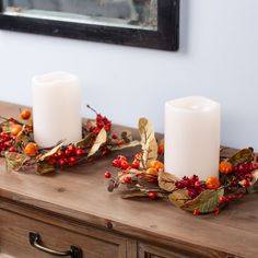 two white candles sitting on top of a wooden table covered in leaves and red berries