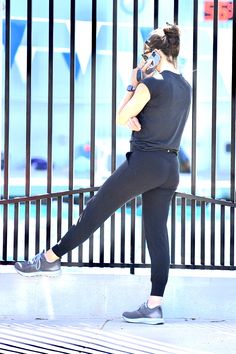 a woman in black is leaning against a fence and talking on her cell phone while wearing running shoes