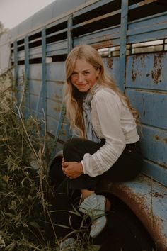 a woman sitting on the side of a blue truck next to tall grass and weeds