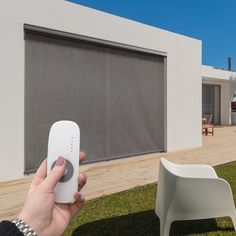 a hand holding a remote control in front of a white chair and window with roller shades