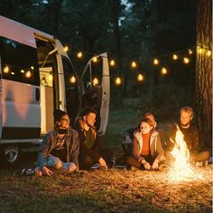 four people sitting around a campfire in front of a van with lights strung over it