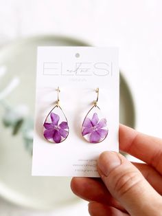 a person holding up a pair of earrings with purple flowers on the front and back