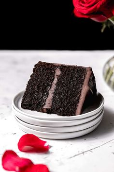 a piece of chocolate cake on a white plate with red rose petals around it and a vase in the background