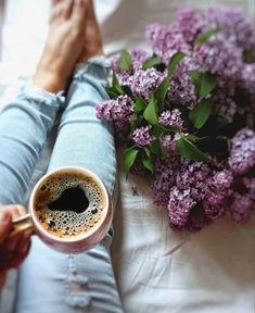 a woman holding a cup of coffee next to purple flowers on a white sheet with her legs crossed