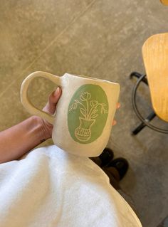 a person holding a coffee cup with a plant on it in their left hand while sitting next to a chair