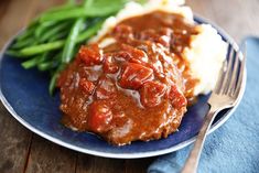 a blue plate topped with meat covered in gravy next to green beans and mashed potatoes