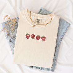 Welcome the sunny days with our delightful Summer Strawberry Embroidered T-Shirt! Made from 100% cotton, this t-shirt is not only comfortable but also perfect for staying cool during warm weather. Available in sizes S to 2XL and nine vibrant colors including Banana, Bay, Blossom, Butter, Chalky Mint, Chambray, Ivory, Orchid, and Peachy, there's a shade to suit every style.The standout feature of this t-shirt is the charming summer fruit-inspired design, showcasing four intricately embroidered strawberries on the center chest. Whether you're heading to a picnic, beach outing, or simply enjoying a leisurely day, this t-shirt adds a touch of sweetness to your summer wardrobe.Please note that colors may vary slightly due to monitor settings. For accurate sizing, refer to the size charts provid Cute Cream Cotton T-shirt, Cute Organic Cotton T-shirt For Everyday, Trendy Embroidered Summer T-shirt, Trendy Summer T-shirt With Embroidered Text, Trendy T-shirt With Embroidered Text For Summer, Trendy Embroidered T-shirt For Summer, White Embroidered T-shirt For Summer, Trendy Embroidered Cotton T-shirt, Summer Everyday T-shirt With Embroidered Text