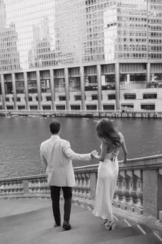 a man and woman holding hands while walking on a bridge over water in front of tall buildings