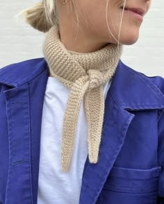 a woman wearing a blue jacket and a beige knitted neck scarf is standing in front of a white brick wall