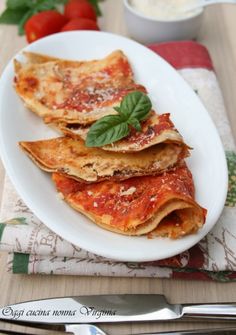 a white plate topped with lasagna covered in sauce and basil next to a bowl of tomatoes