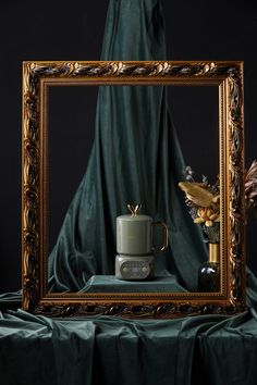 a gold frame with a coffee cup and vase in front of it on a green cloth