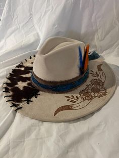 Off white wide brim, Cow horns and flowers burnt on brim. Cowhide material with blue band and feathers. White Western Felt Hat For The Beach, White Hat Bands For Spring Ranch, White Hat Band For Spring Ranch Occasions, White Brimmed Fedora For Festival, Spring White Hat Band For Ranch, White Fedora Hat For Festival, Custom White Fedora For Country Events, White Fedora With Short Brim For Festivals, Country Style White Felt Hat For The Beach