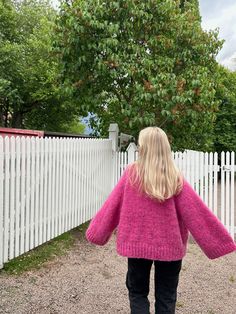 a woman standing in front of a white fence wearing a pink sweater and black pants