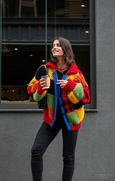 a woman standing on the sidewalk holding a cup and wearing a colorful knitted jacket