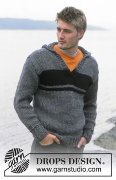 a man standing next to the water wearing a gray and black sweater with an orange shirt